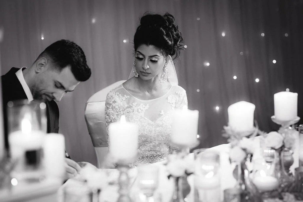 Black and white photo of a bride and groom at their wedding ceremony, with the couple looking down, surrounded by candles and floral arrangements.