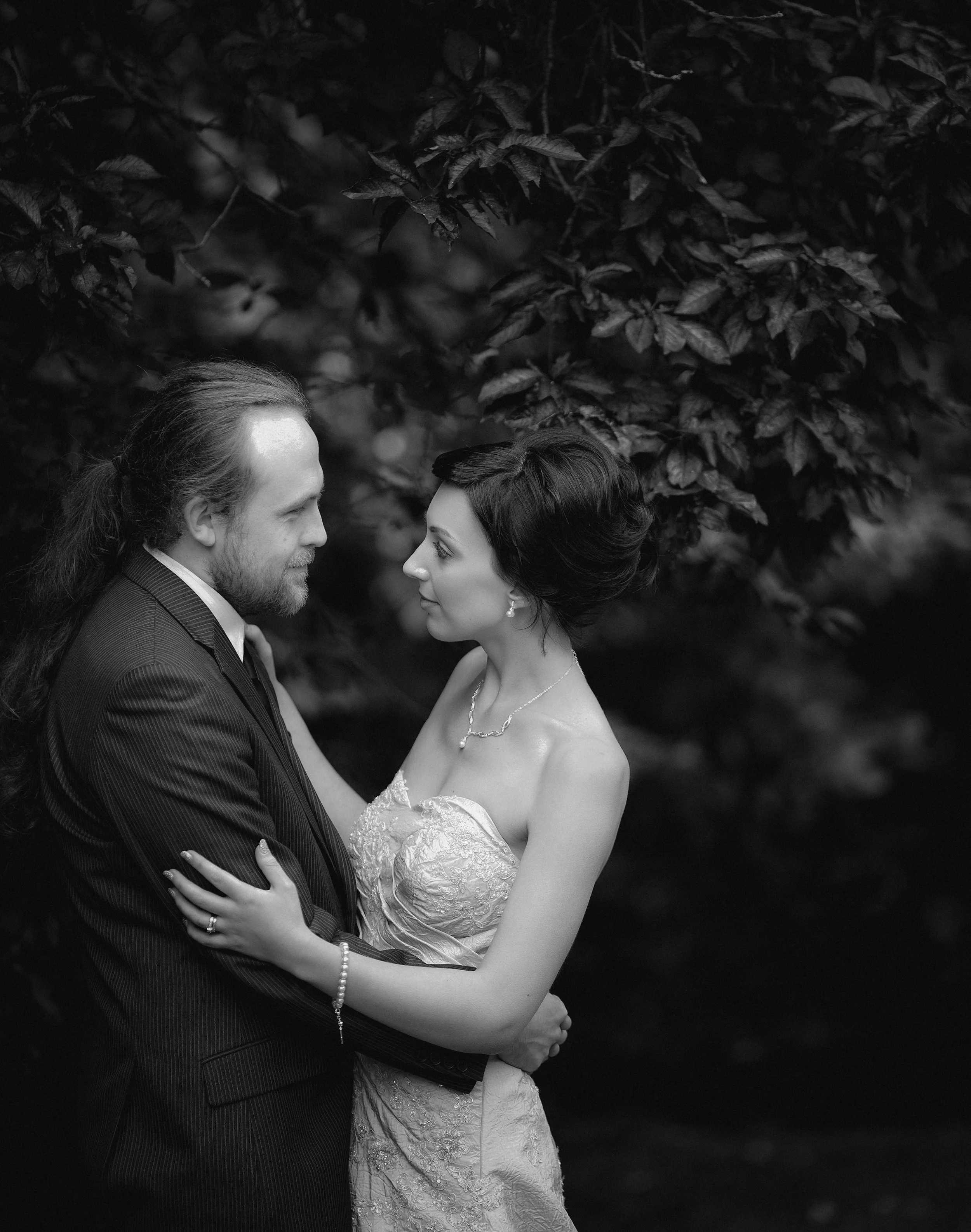 A black and white photo of a bride and groom gazing into each other's eyes outdoors, surrounded by trees and foliage.