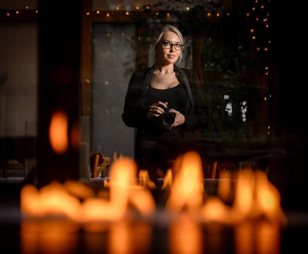 Woman with glasses holding a camera in a dimly lit room, with reflected candles and string lights creating a warm atmosphere.