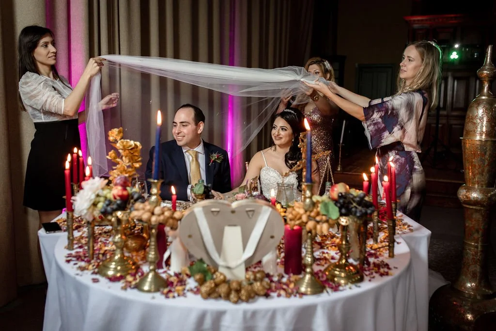 A newlywed couple sits at a decorated wedding reception table with candles, flowers, and food, while friends lift a sheer cloth over them in a celebration moment.