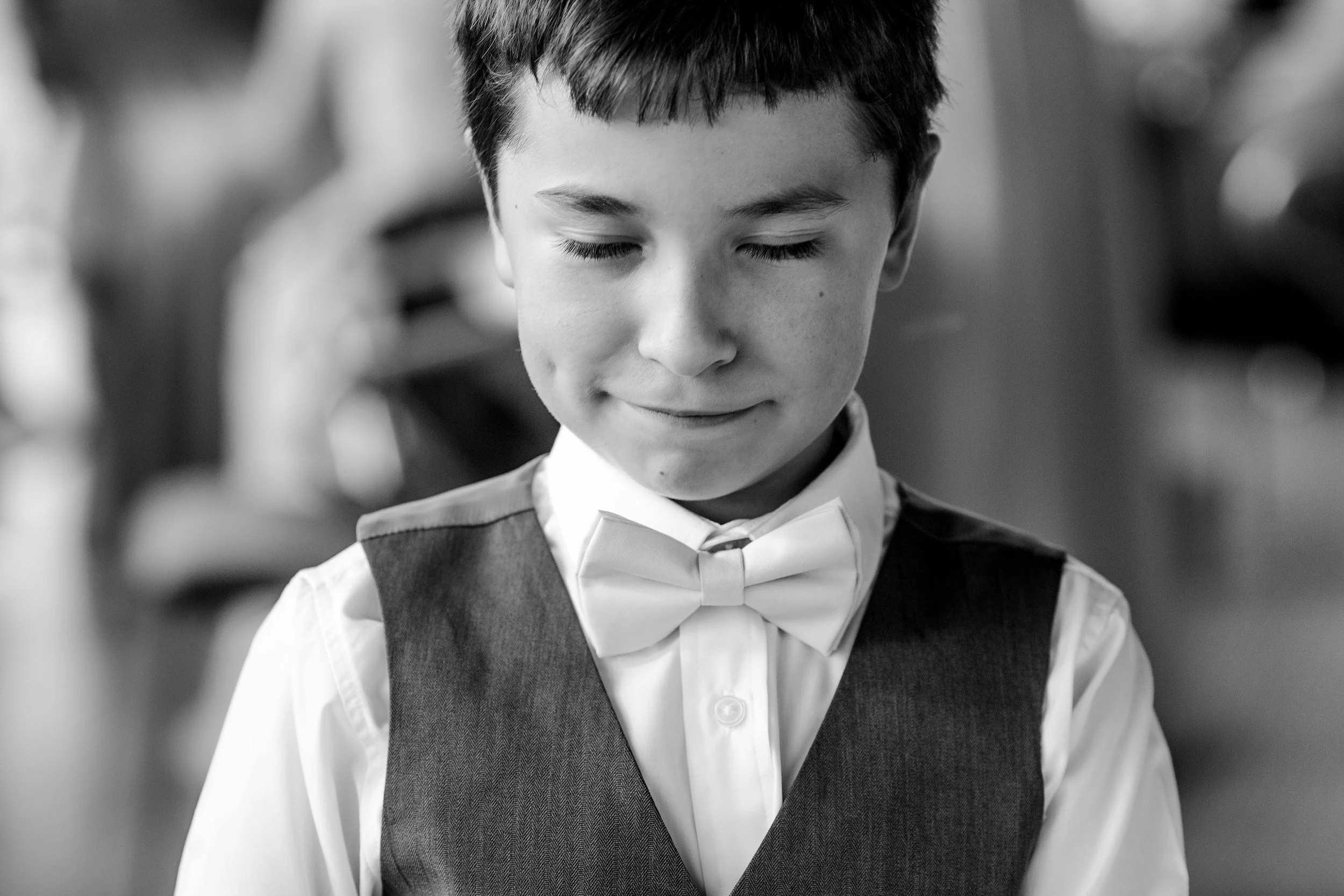 A young boy with closed eyes, dressed in a white shirt, gray vest, and bow tie, smiling softly.