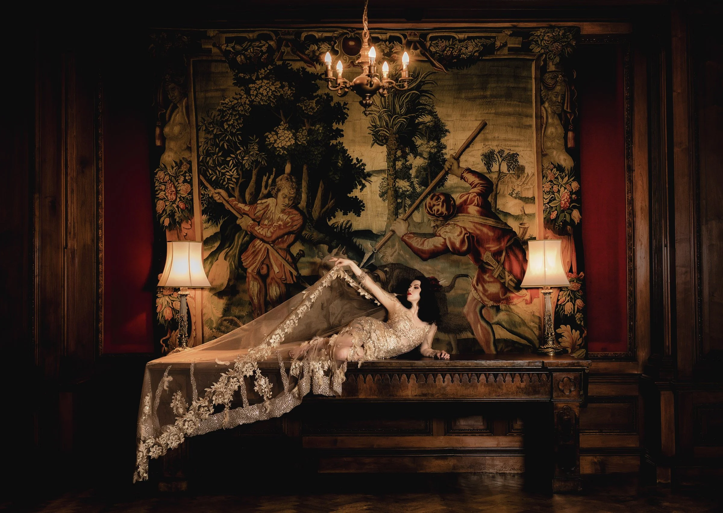 A woman in an ornate gold lace gown lying on a wooden table in a dimly lit room, with a large tapestry of two men fighting with spears behind her, flanked by two lit lamps and a chandelier above.