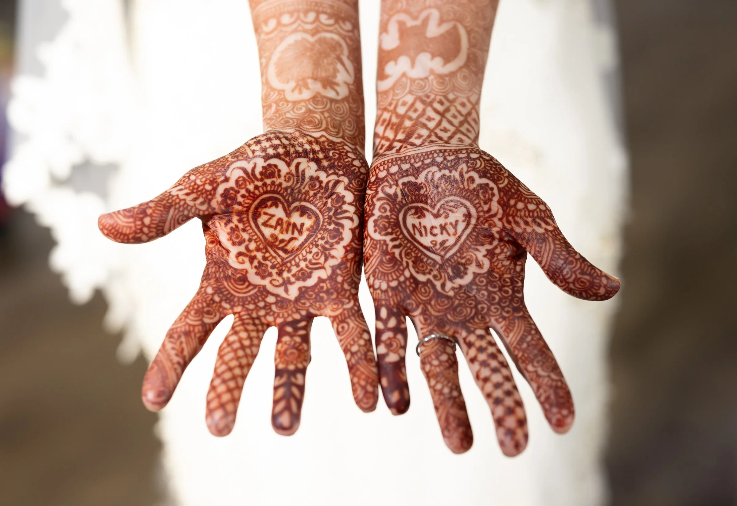 Two hands with intricate henna designs, each with a heart-shaped name inside. The left hand's heart reads 'Zain' and the right hand's reads 'Nicky'.