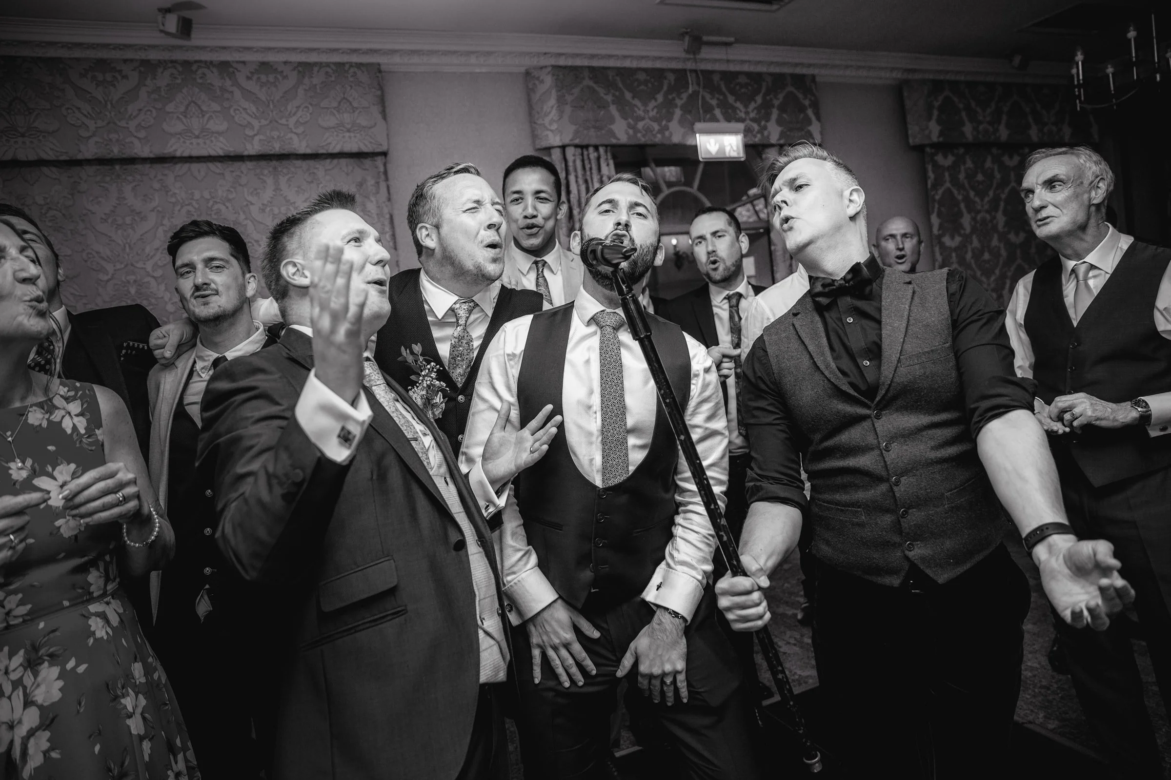 Group of men and women singing and celebrating at an indoor event, dressed in formal attire, with a microphone stand in front.