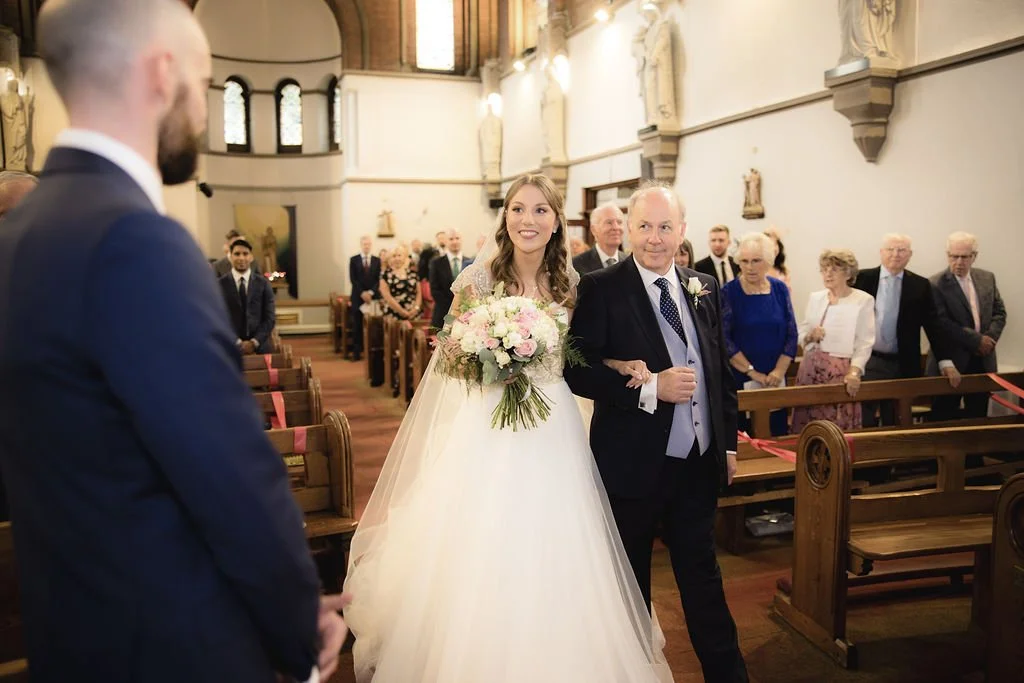 A bride walking down the aisle with her father in a church, with guests seated and watching. The bride is holding a bouquet of flowers and smiling.