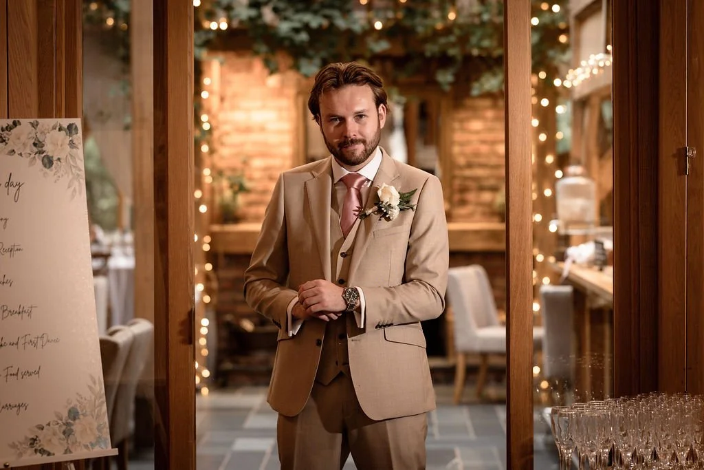 A man in a beige suit with a white boutonniere stands inside a decorated venue, possibly for a wedding, with warm string lights and wood-paneled walls.