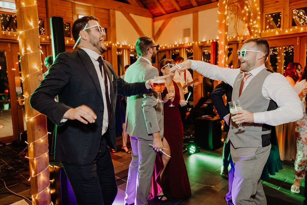 People dancing and having fun at a party or celebration, wearing sunglasses with humorous designs, in a decorated wooden venue with string lights.
