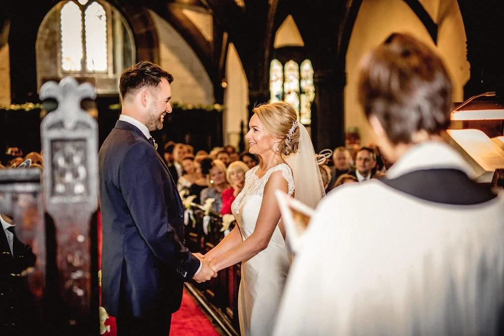 A bride and groom holding hands and smiling at each other during their wedding ceremony inside a church, with guests seated and a priest officiating.