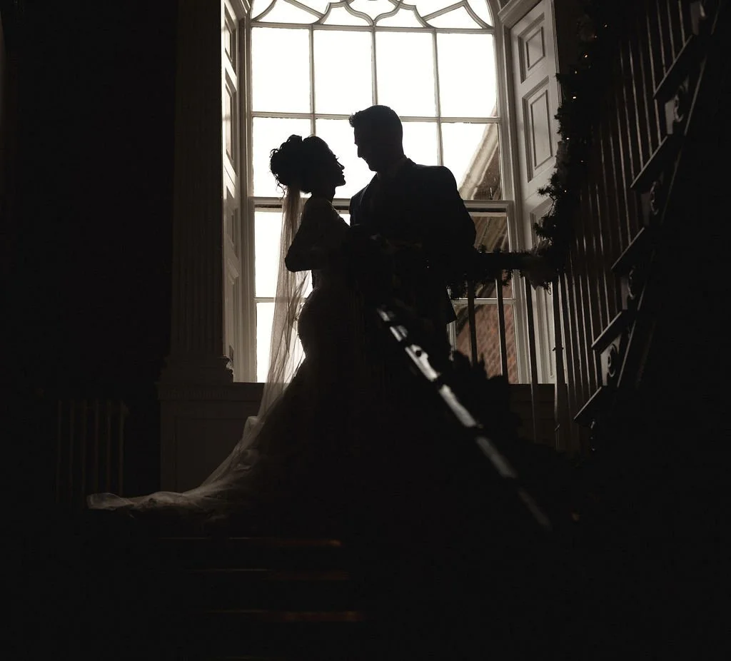 Silhouettes of a bride and groom standing close together on a staircase in front of a large window with daylight streaming in.