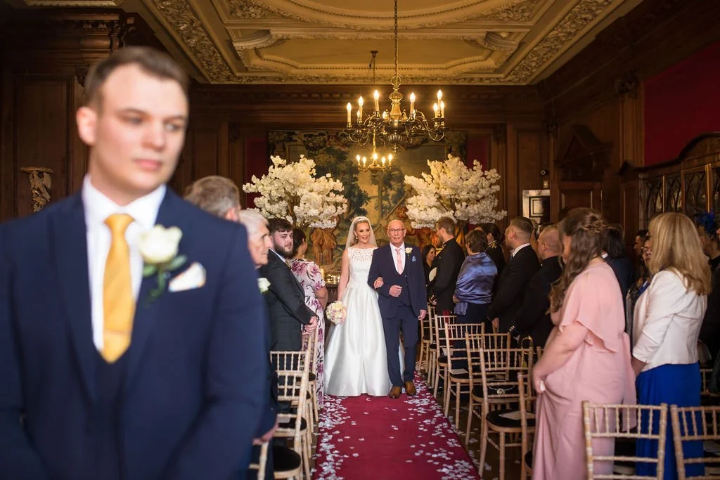 A wedding ceremony taking place in an elegant room with wood-paneled walls, ornate ceiling, and a chandelier. The bride, in a white wedding dress, walks down the aisle escorted by a man in a navy suit. Guests are seated on gold chairs, facing the ais