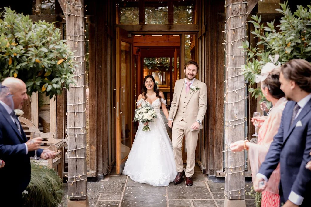 A newly married couple walking out of a rustic wooden venue, smiling, as guests celebrate and raise glasses with confetti and flowers.