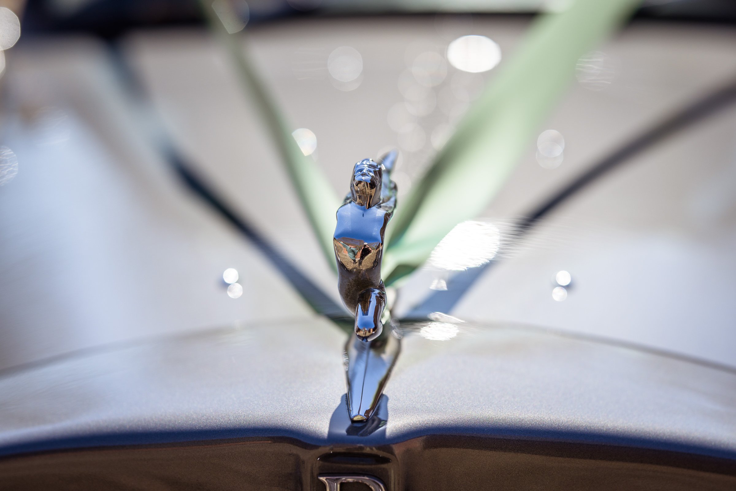 Close-up of a vintage car hood ornament, a metallic sculpture of a woman with outstretched arms, mounted on the hood of a gray car, with green plant leaves and blurred bokeh lights in the background.
