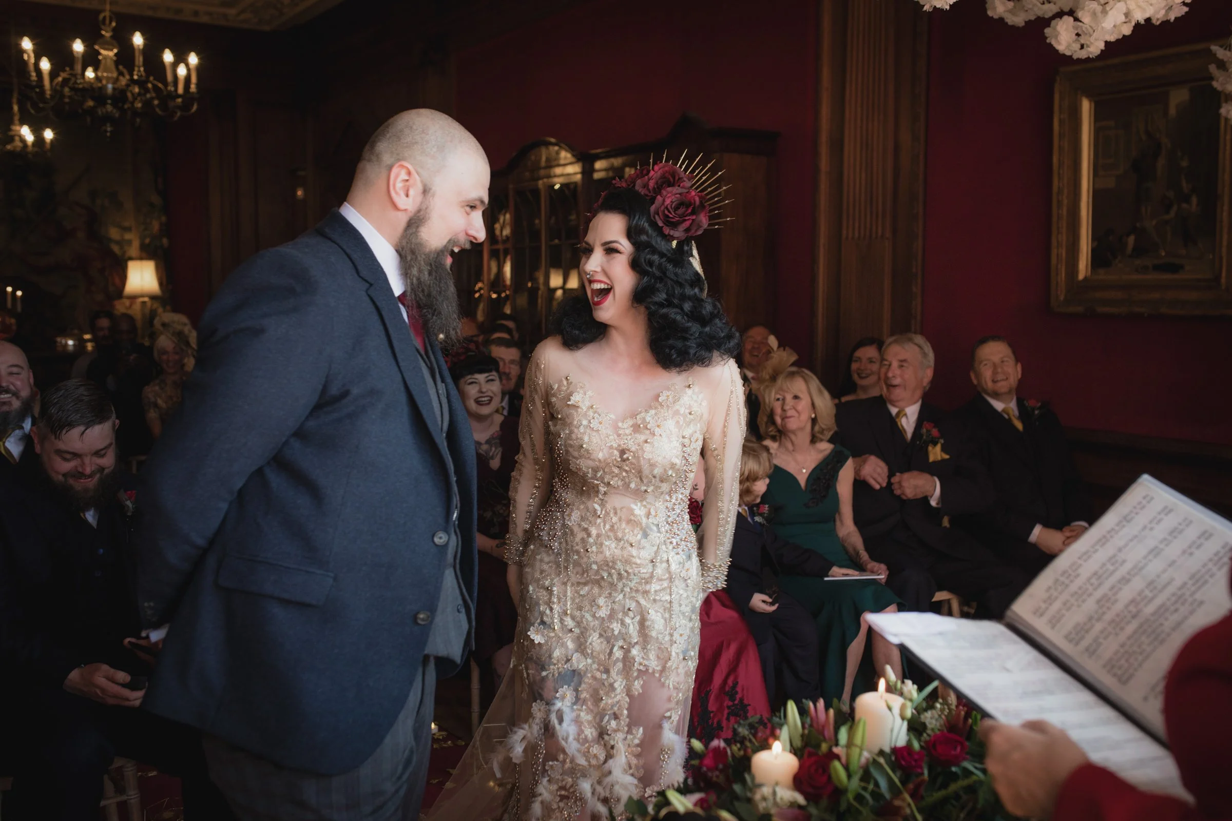 Bride and groom exchanging vows during wedding ceremony with guests smiling and laughing in the background, decorated with candles and flowers