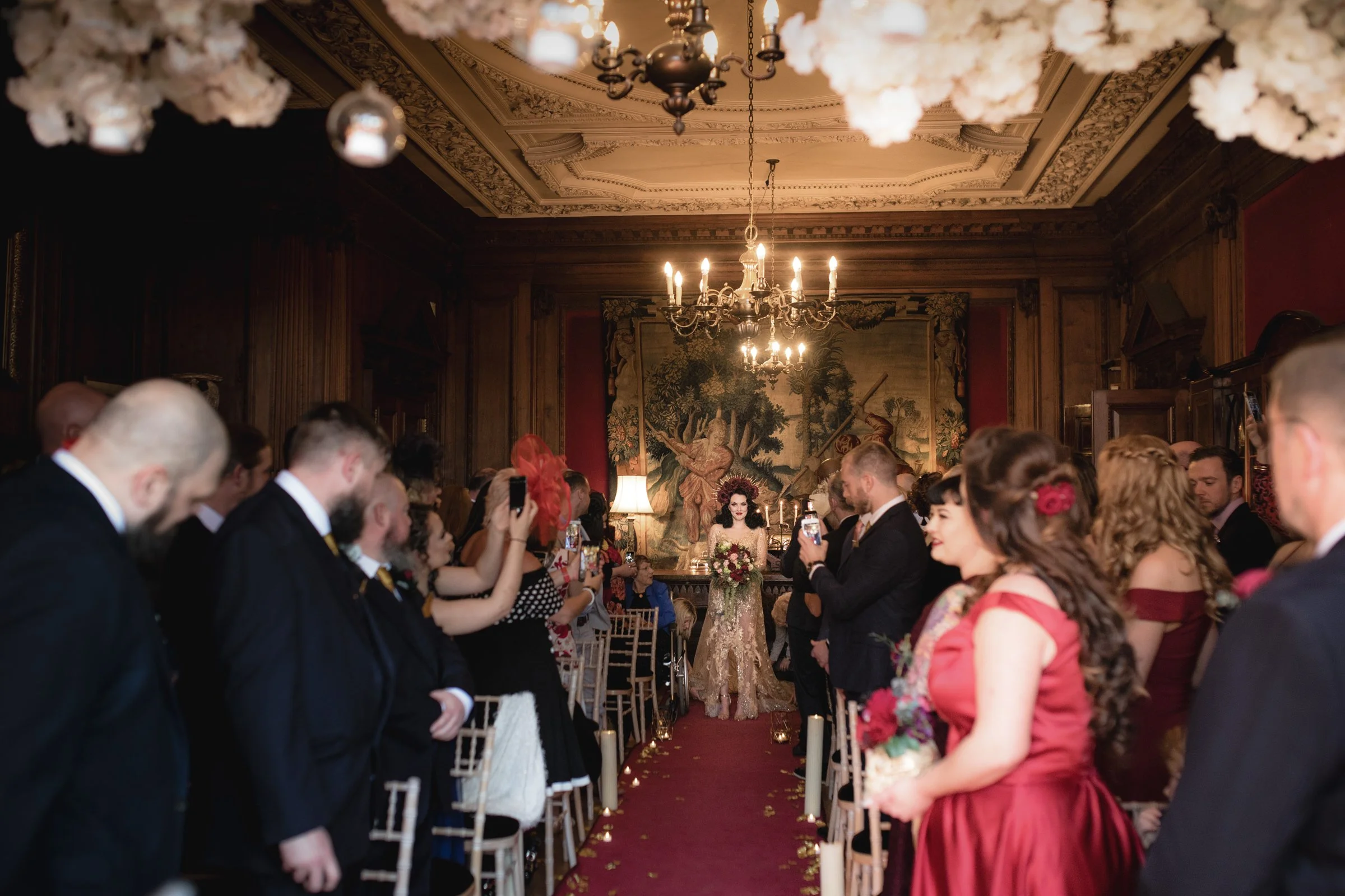 Bride in a gold lace dress holding a bouquet, dressed in vintage style, standing at the end of an aisle in a richly decorated room with dark wood paneling, chandeliers, large tapestry on the wall, and guests standing and taking pictures.