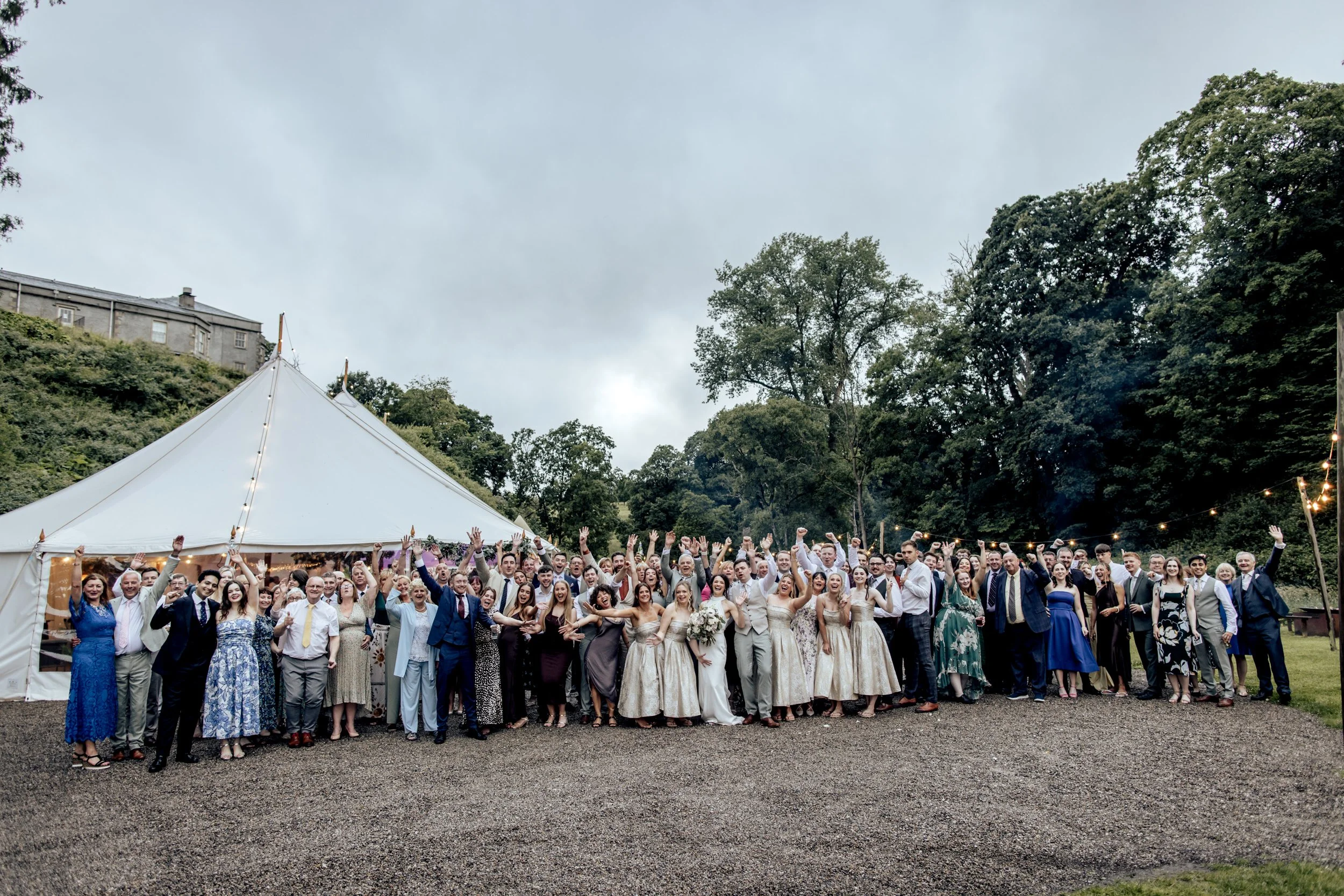 Large group of wedding guests gathered outside near a white tent, celebrating under cloudy skies surrounded by trees and a hillside.