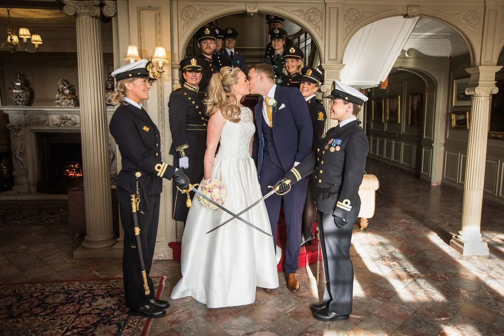 Bride and groom share a kiss while surrounded by women in naval uniforms in a grand, elegantly decorated room.