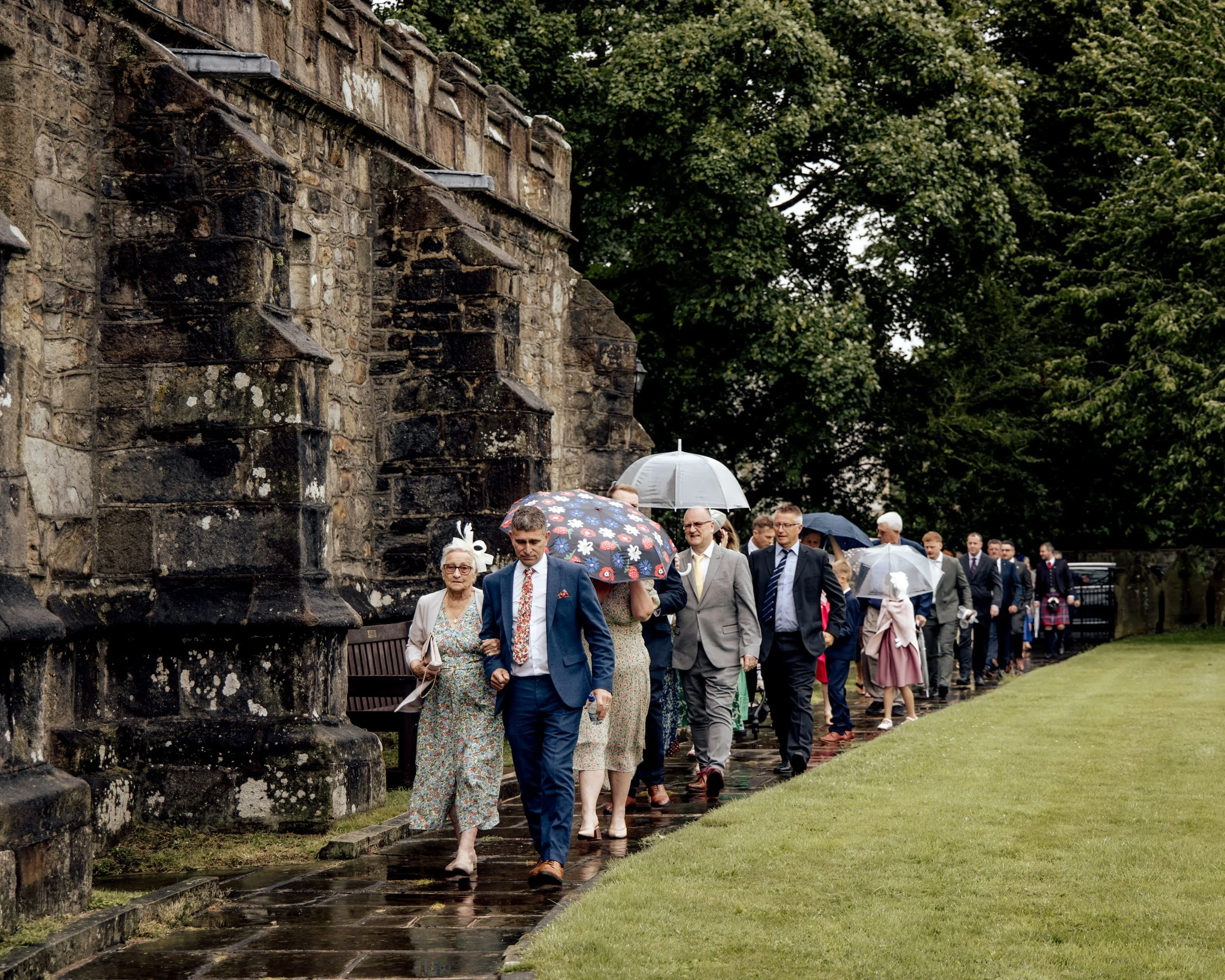 People dressed in formal attire walking on a paved path beside an old stone wall, holding umbrellas on a rainy day, with green trees in the background.