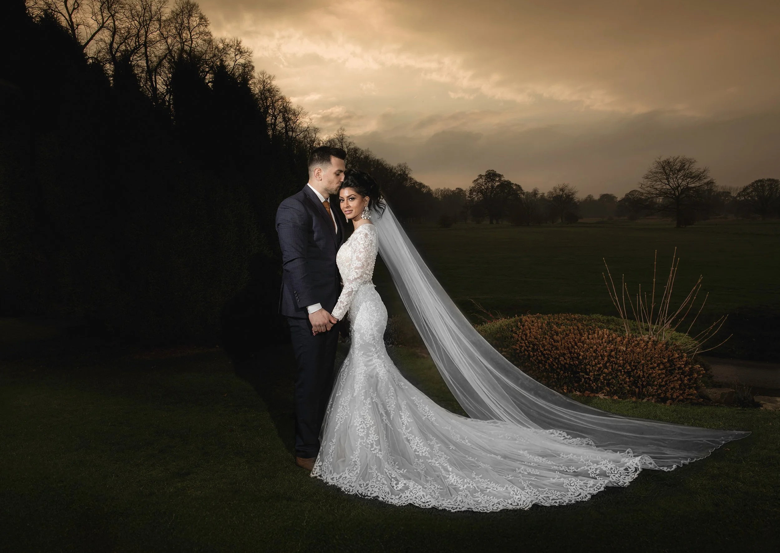 Bride and groom holding hands outdoors at sunset, bride in a white lace wedding gown with a long train and veil, groom in a dark suit, trees in the background.