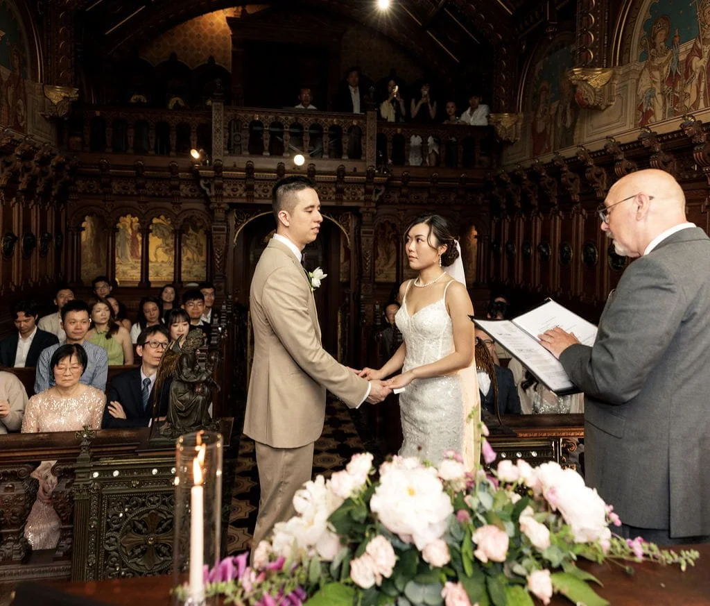 A wedding ceremony taking place in a church with a bride and groom holding hands in front of an officiant. The bride wears a white dress and the groom wears a beige suit. Guests are seated and watching, some with expressions of emotion. The church fe