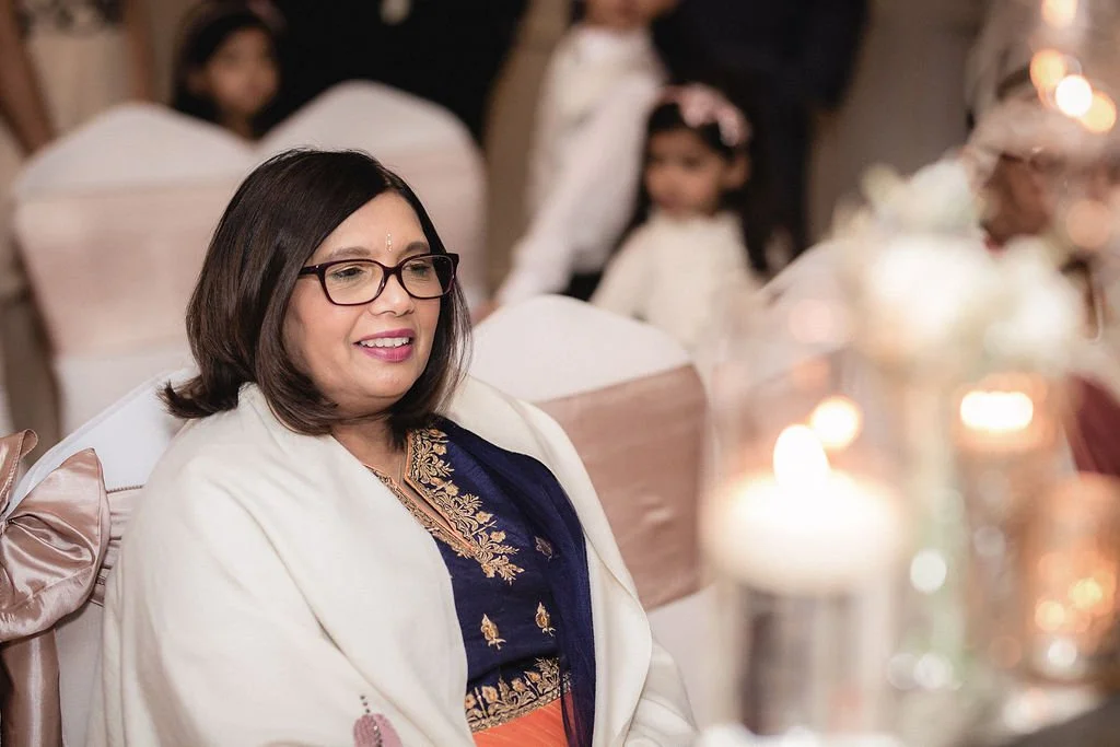 A woman with glasses and shoulder-length dark hair smiling at a formal event, sitting at a decorated table with blurred candles and flowers in the foreground. Behind her are children dressed in white.