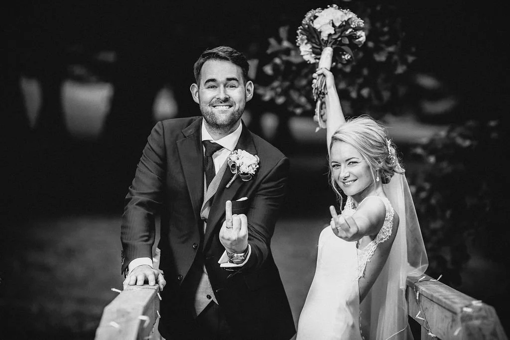 Black and white photo of a smiling bride and groom making hand gestures. The groom, in a suit, stands on the left, resting his hand on a wooden railing. The bride, in a wedding dress with lace details and a veil, stands on the right, raising a bouque