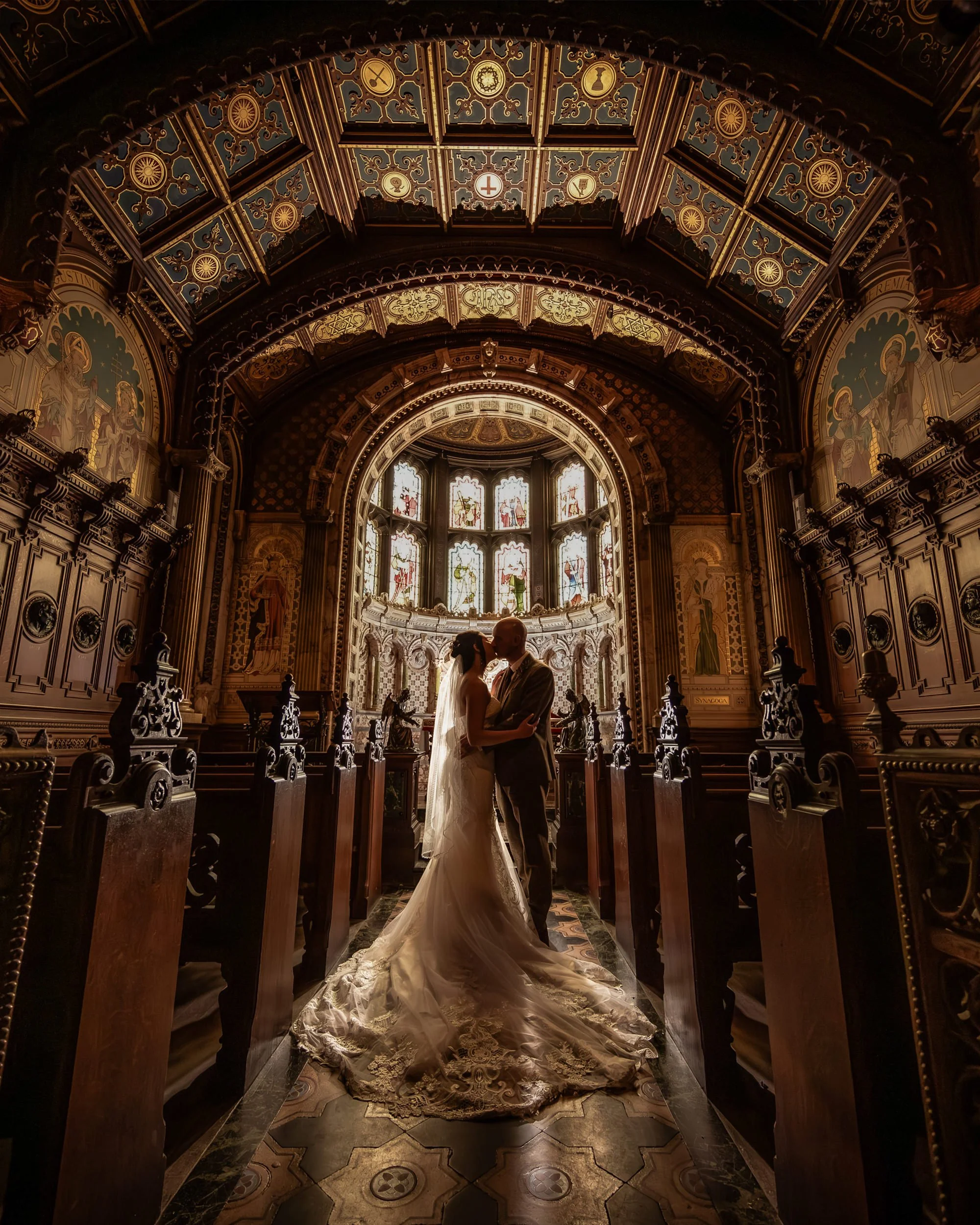A bride and groom embrace in a church with ornate decorations, stained glass windows, and a high, decorated ceiling.