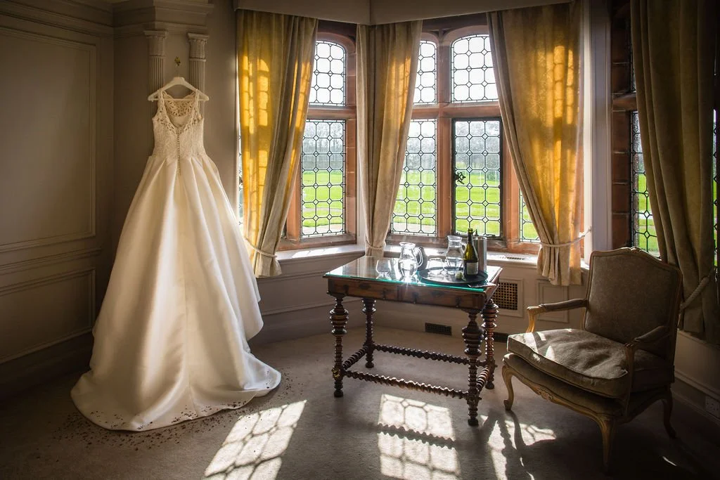 A wedding dress hangs on a wall, illuminated by natural sunlight through a large window with leaded glass panes in a warmly lit room with cream-colored curtains and vintage furniture.
