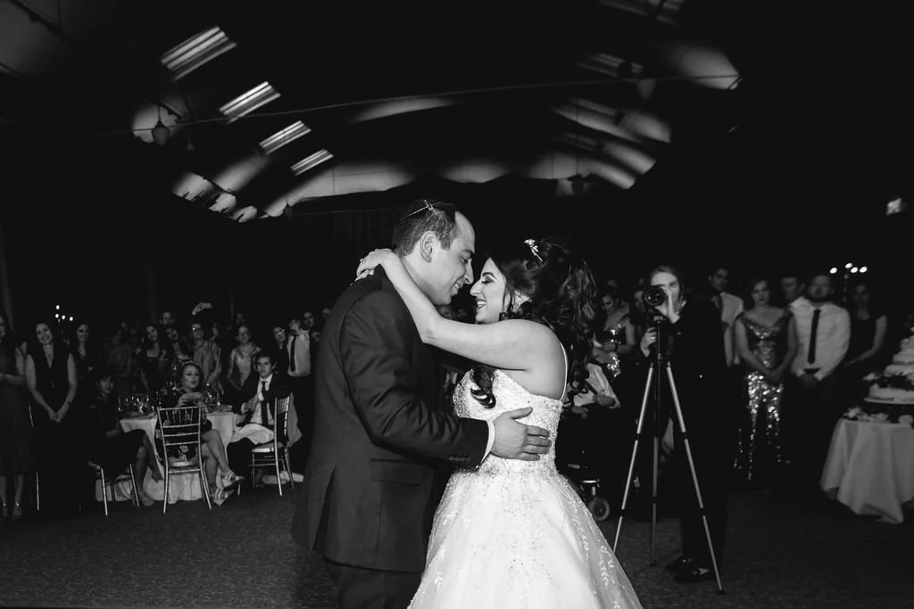 Couple dancing at wedding reception, surrounded by guests, in a large hall with high ceiling and bright lights, black and white photo.