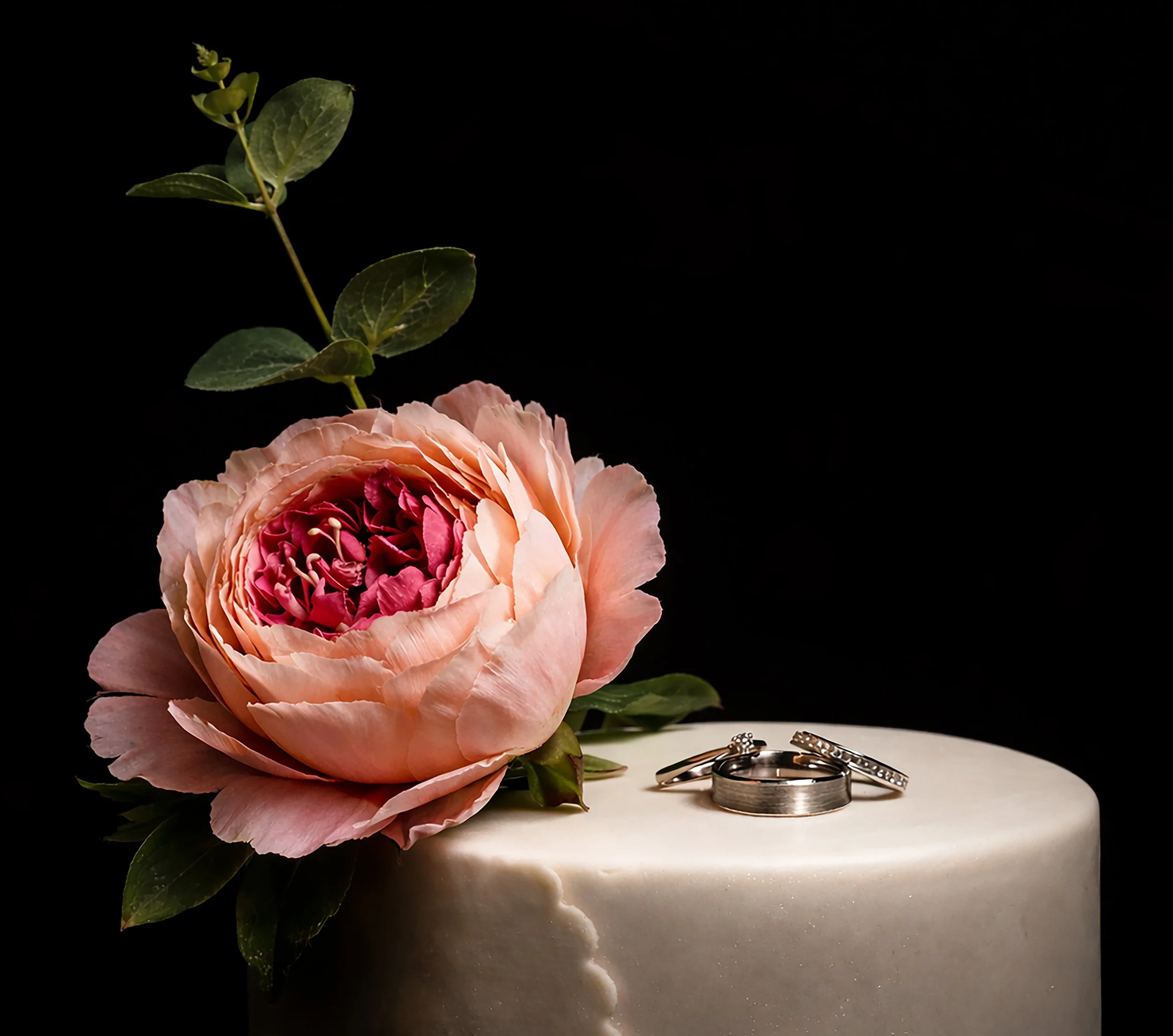 A wedding cake decorated with a pink peony flower and three wedding rings on top, set against a black background.