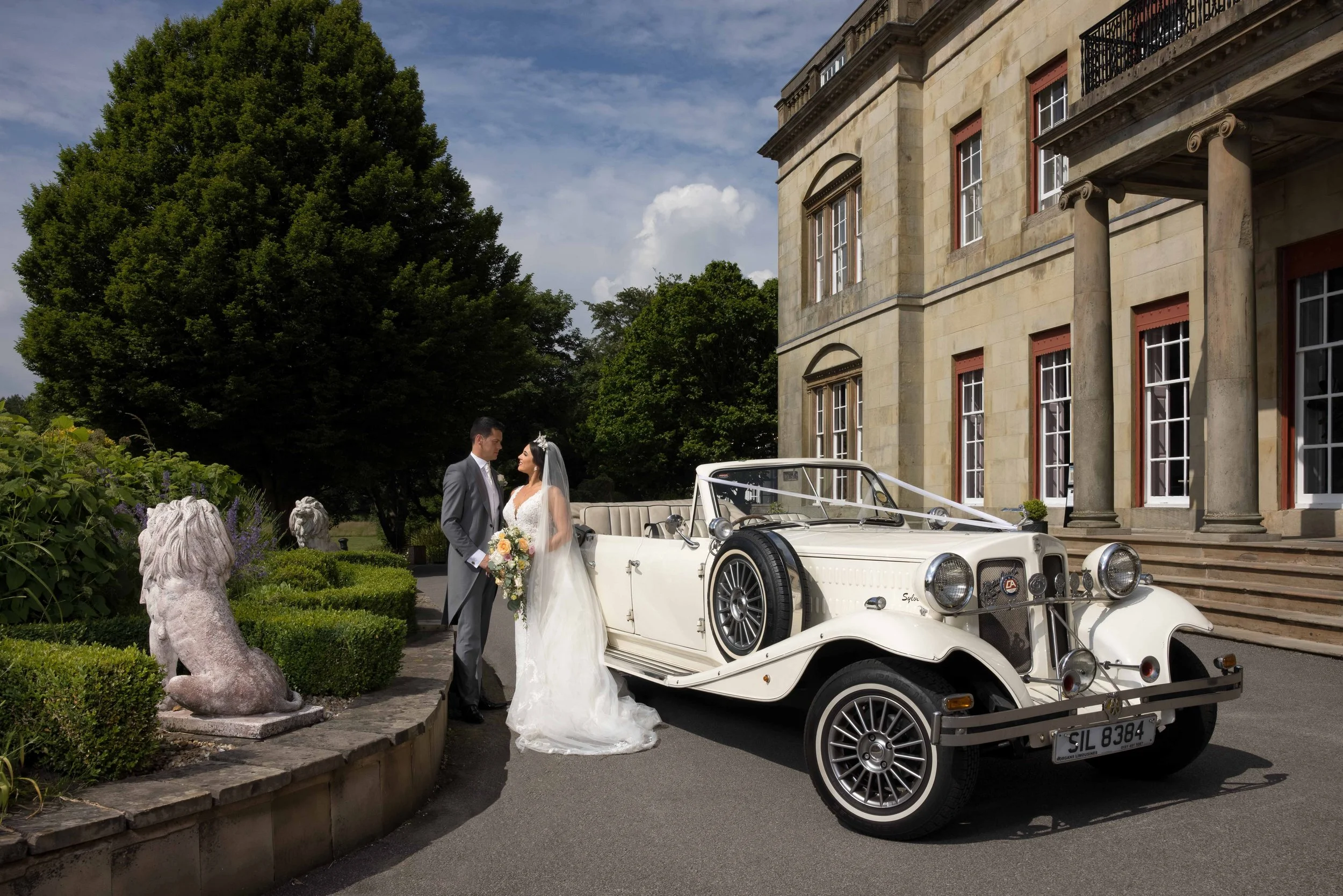 A bride and groom standing next to a vintage white convertible car in front of a large, elegant stone building with columns and multiple windows. The bride is holding a bouquet, and they are looking at each other. Green trees and shrubs surround the 