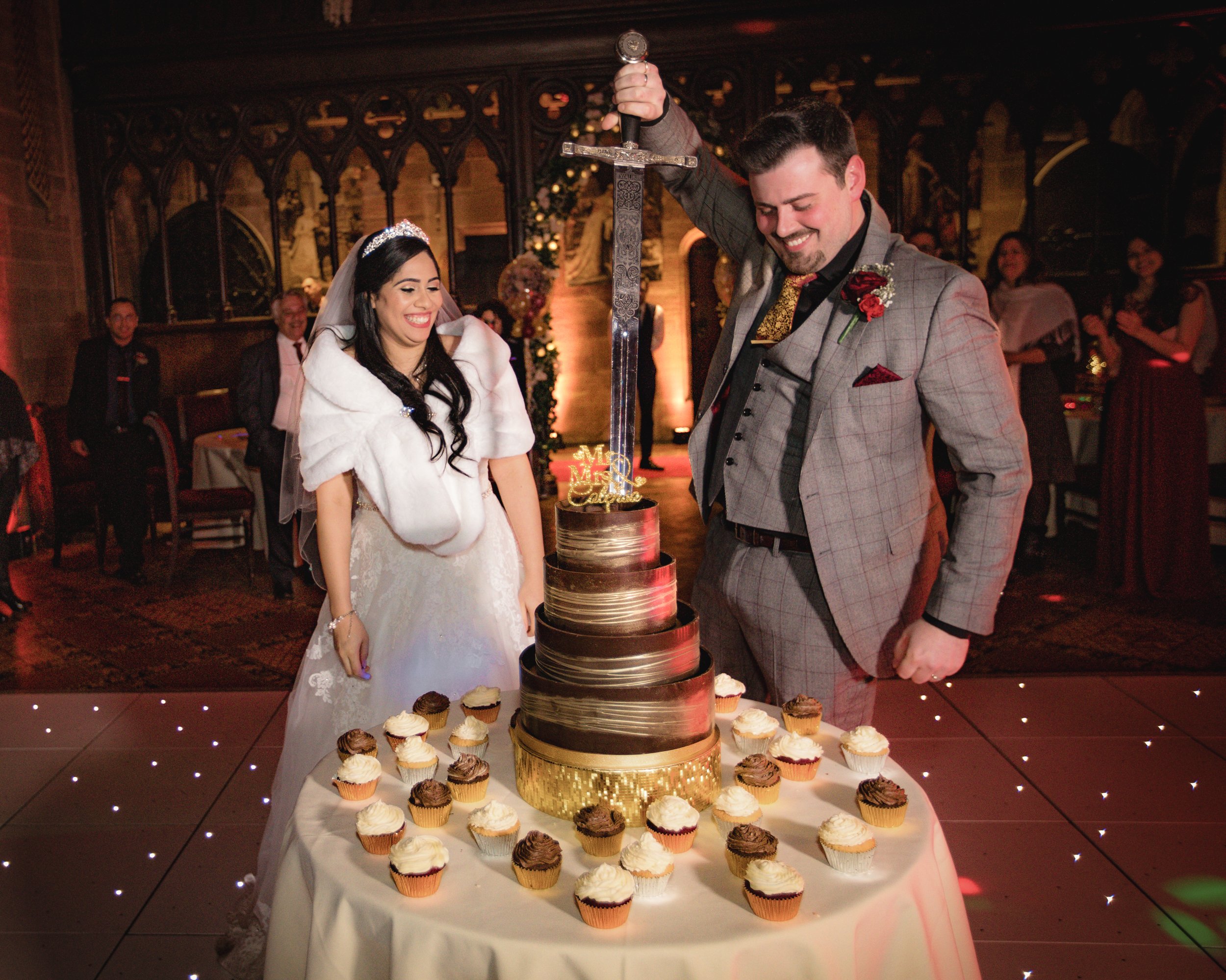 A bride and groom cutting a wedding cake at their reception, surrounded by cupcakes and guests in a decorated venue.
