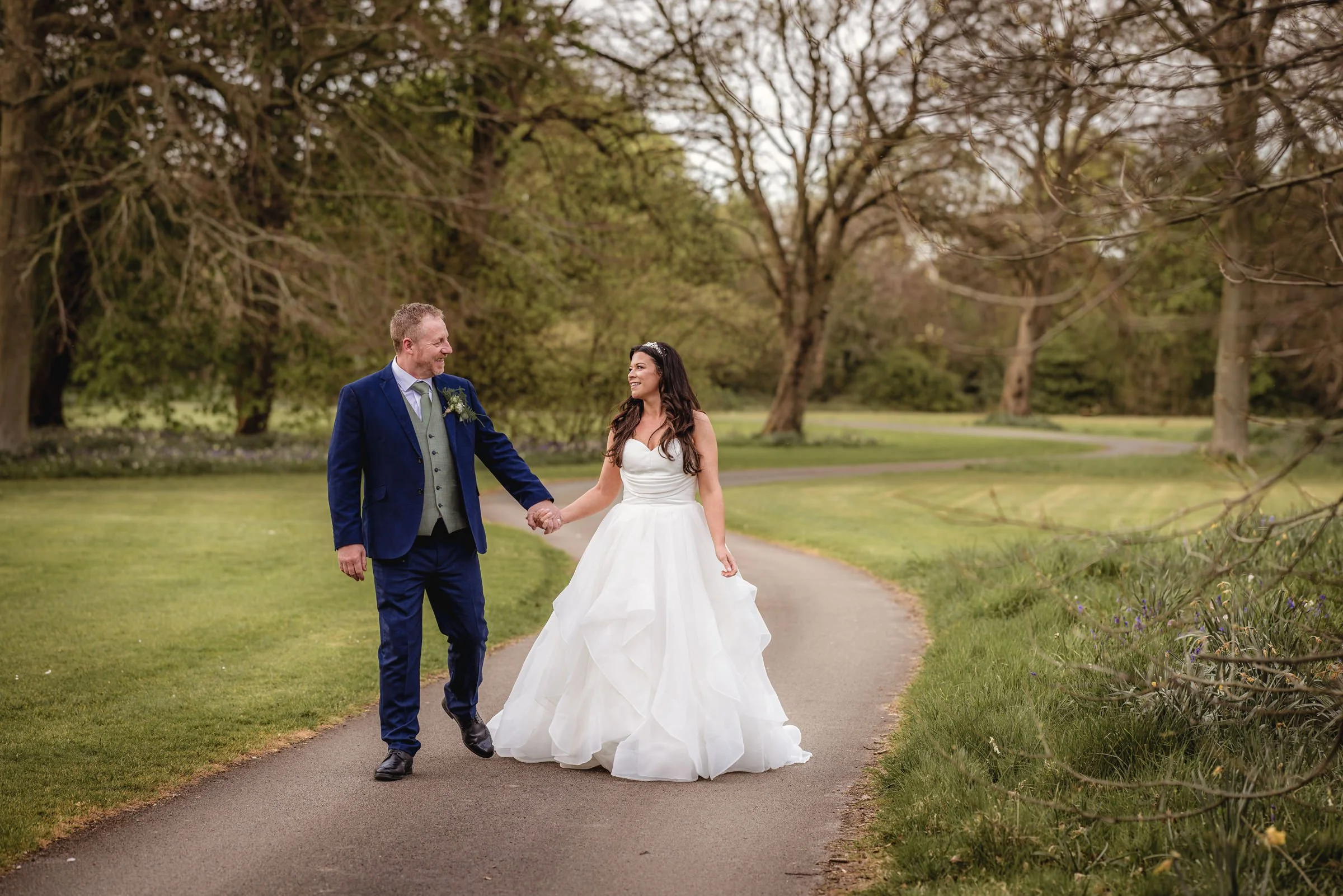 A newlywed couple holding hands and walking on a park path surrounded by trees and grass.