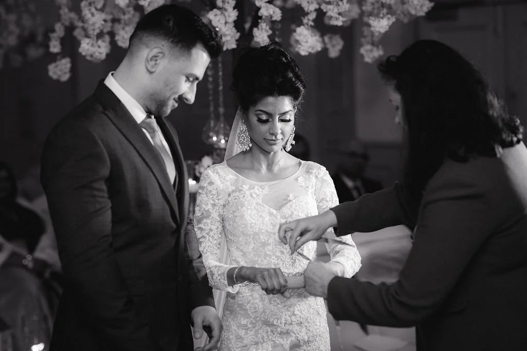 A black and white photo of a wedding ceremony where a woman in a lace wedding dress is being handed a ring by a woman in a dark suit, with a man in a suit standing next to her, all under floral decorations.