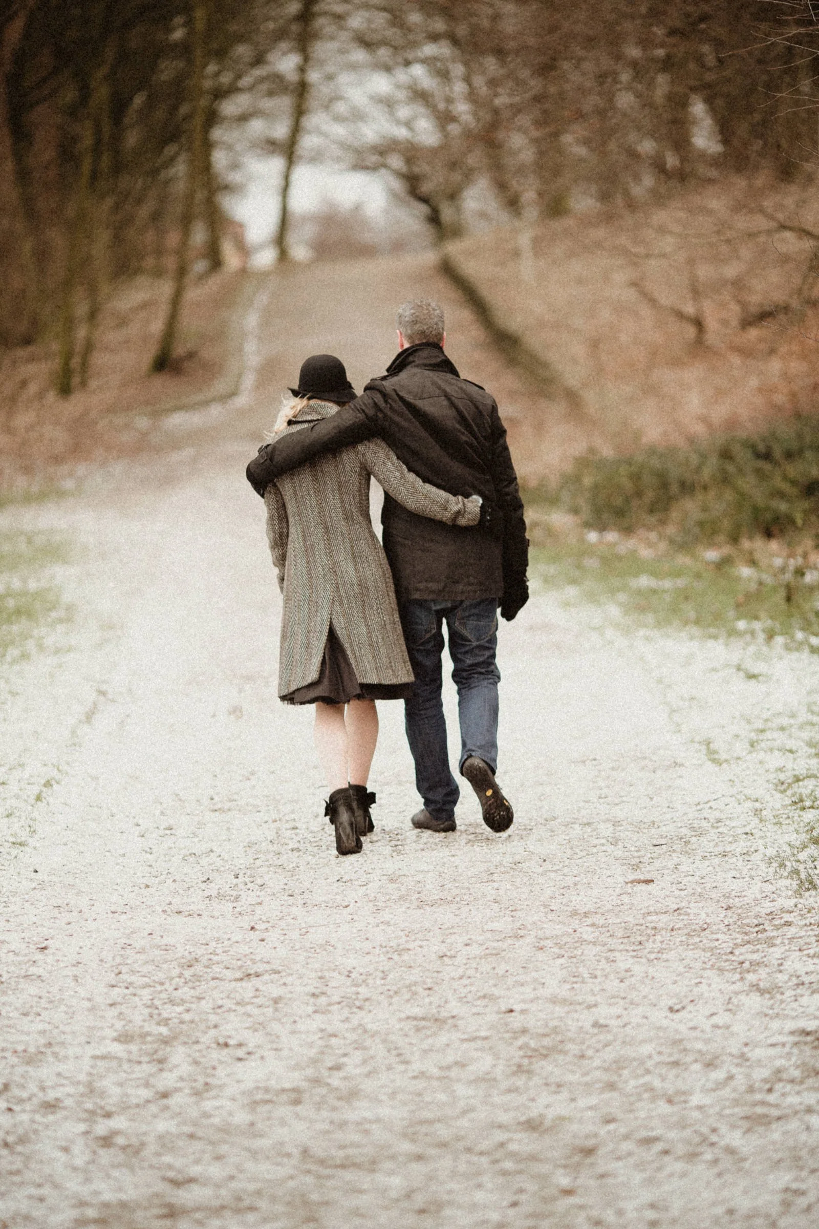 A couple walking arm-in-arm on a dirt path in a park during late autumn or winter.