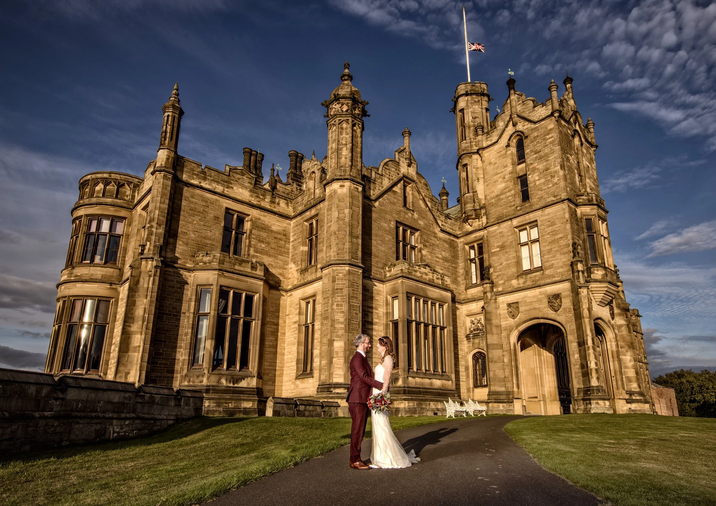A bride and groom holding hands in front of a large castle-like building with a flag flying above it, under a partly cloudy sky.