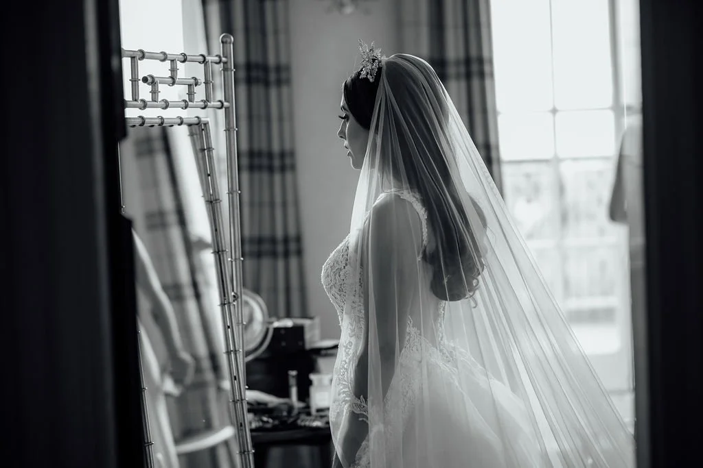 Black and white photo of a bride in lace wedding dress and veil, standing in front of a mirror, indoors near a window.