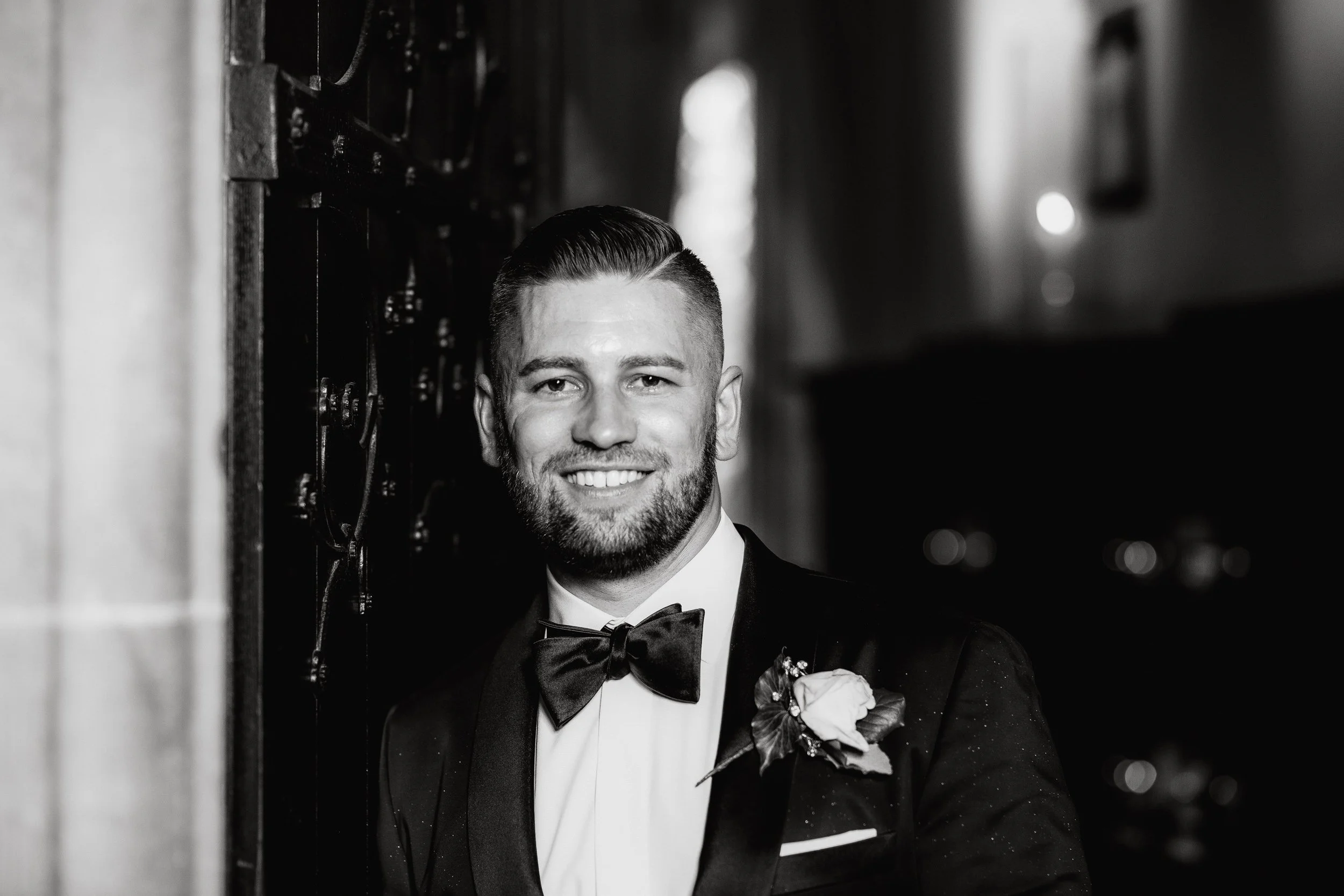 A smiling man in a tuxedo with a bow tie and boutonniere, standing indoors next to a decorated wall.