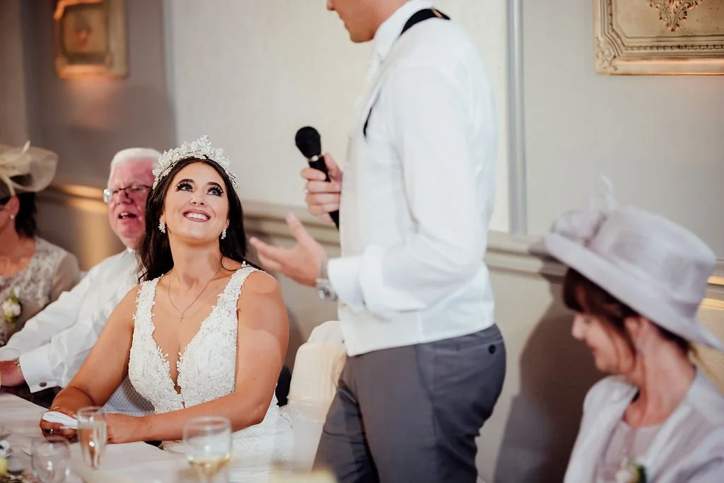 A bride sitting at a wedding reception, smiling and looking up as a man in a white shirt and gray pants gives a speech, holding a microphone. An older man with white hair and glasses, and a woman in a hat are sitting at the table beside her.
