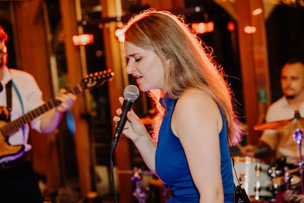 A woman singing into a microphone at a live music venue with guitar and drums in the background.