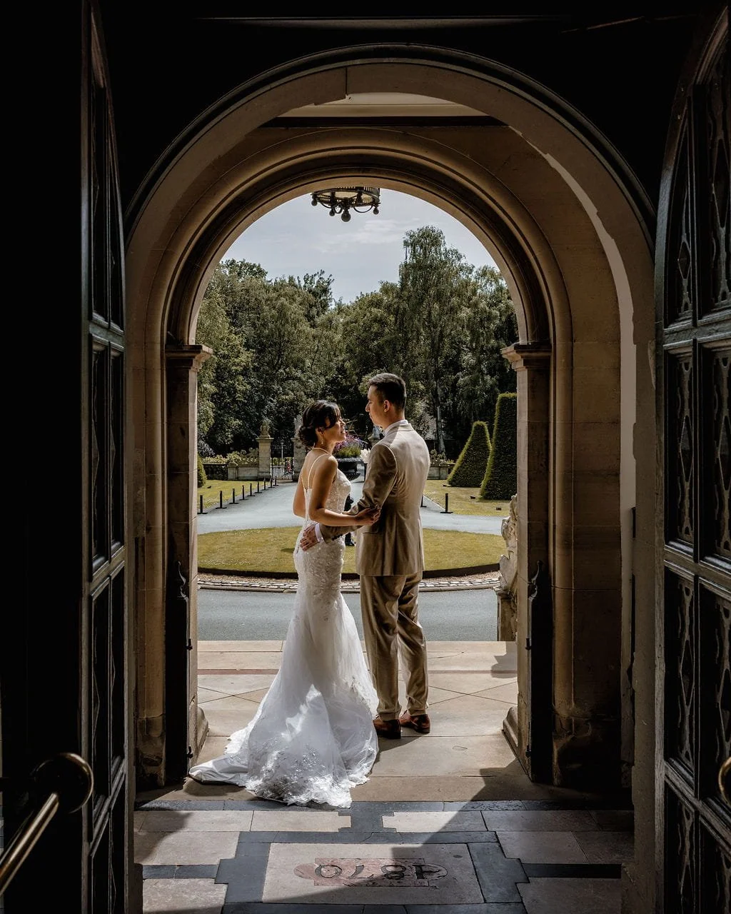 A bride and groom stand holding hands outside a building, framed by an arched doorway, with a scenic garden in the background.
