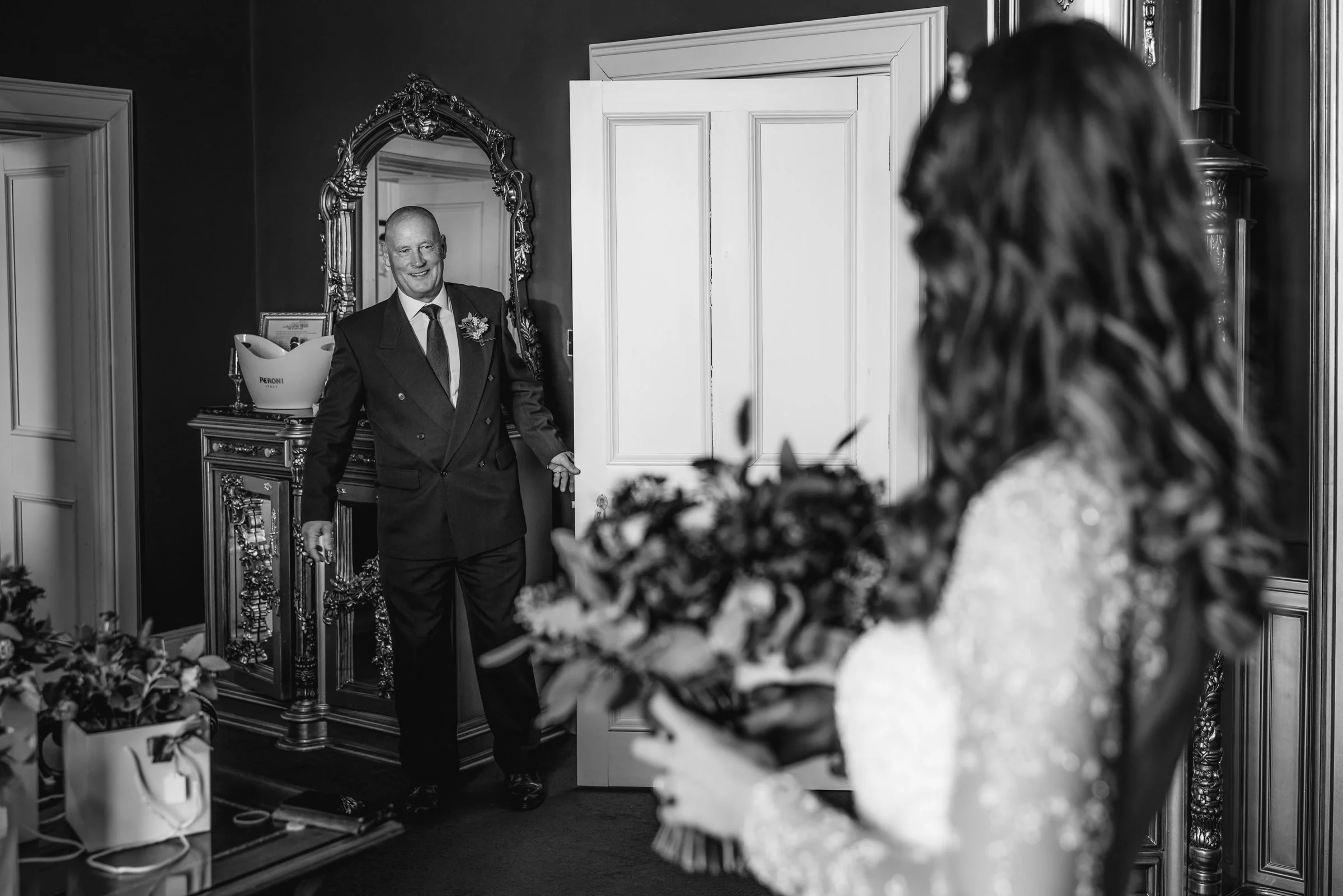 A man in a suit smiling as he enters a room where a woman in a wedding dress holding a bouquet is waiting.