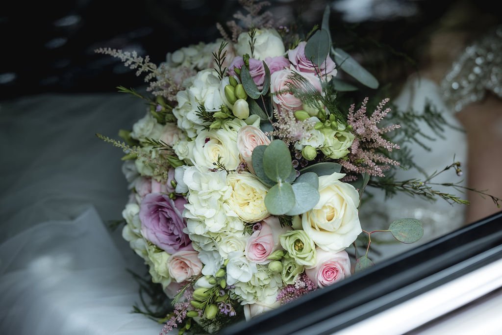 A bridal bouquet of white, pink, and lavender flowers with greenery, resting on a white fabric and glass window.