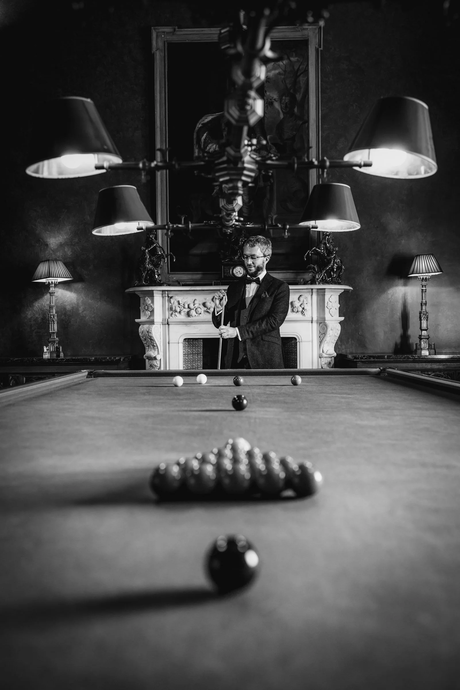 A man in a tuxedo playing billiards in a vintage game room with ornate fireplace, framed painting, and multiple lamps in black and white photography.