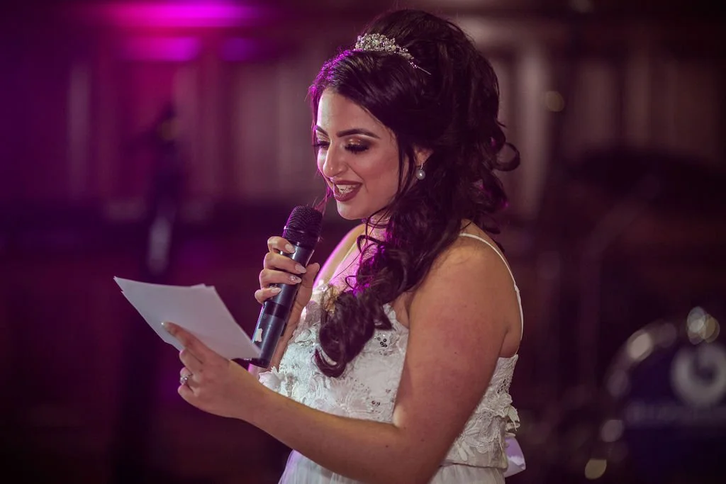 A woman wearing a tiara and a white dress, holding a microphone and a piece of paper, appears to be speaking or singing at an event with dim lighting.