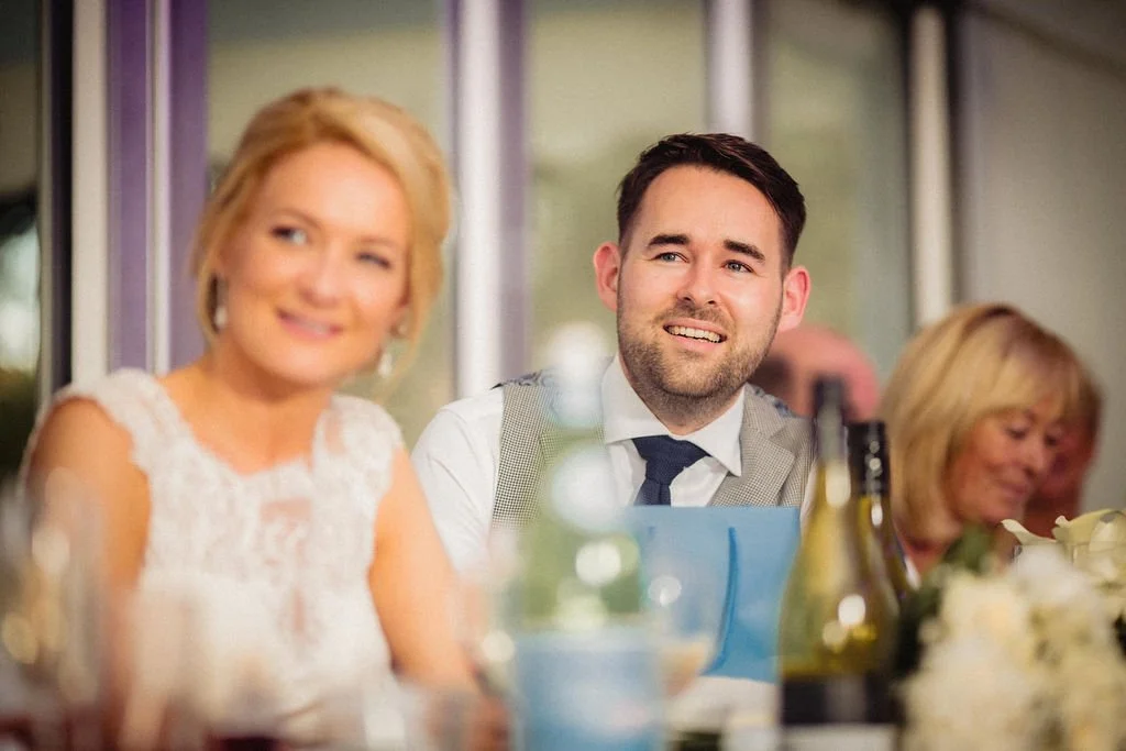 People sitting at a table during a social event, with one woman in a white dress in the foreground, a man in a suit with a vest in the middle, and an older woman in the background.