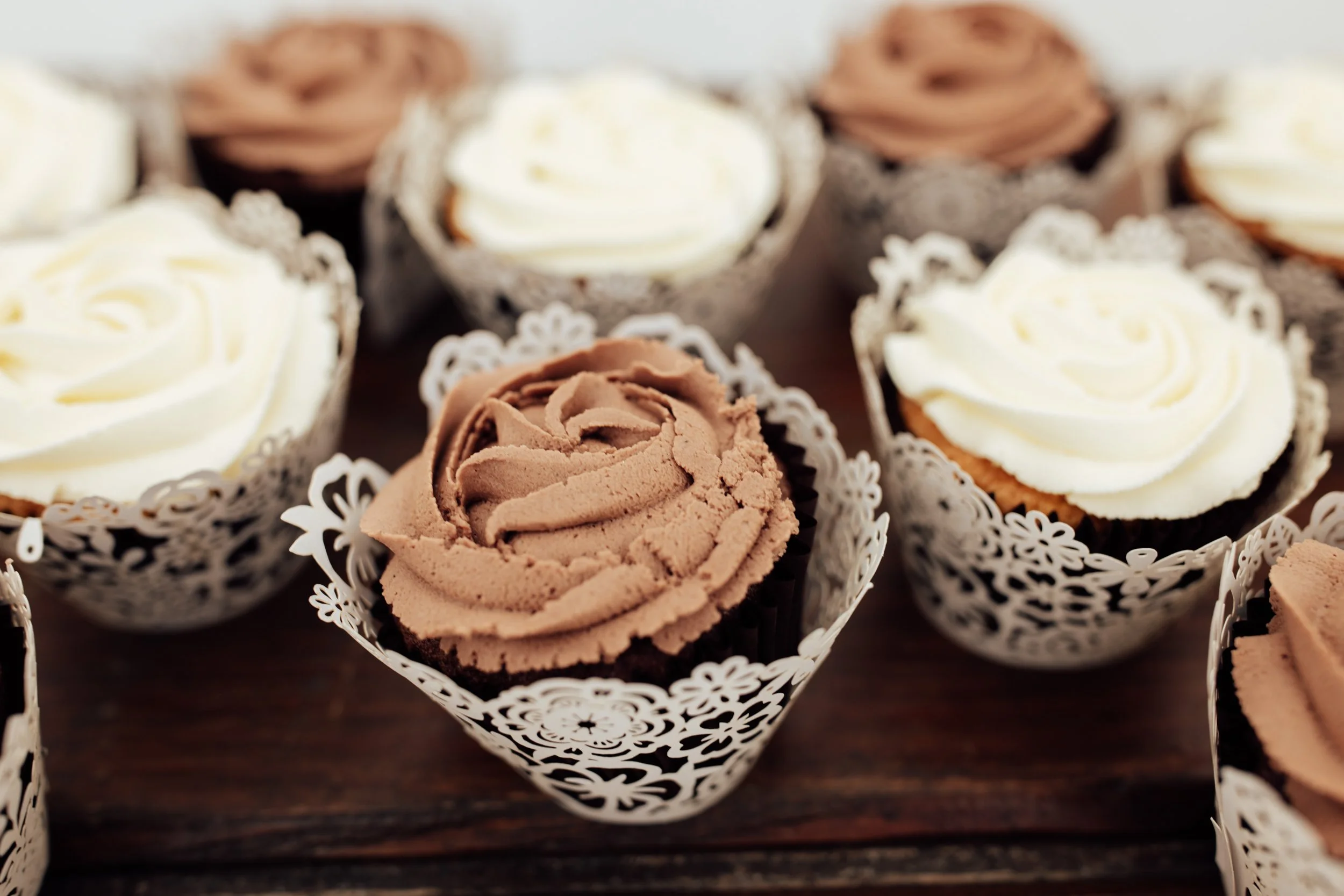 Close-up of chocolate and vanilla cupcakes with swirled frosting, decorated with lace-like paper wrappers, placed on a dark wooden surface.