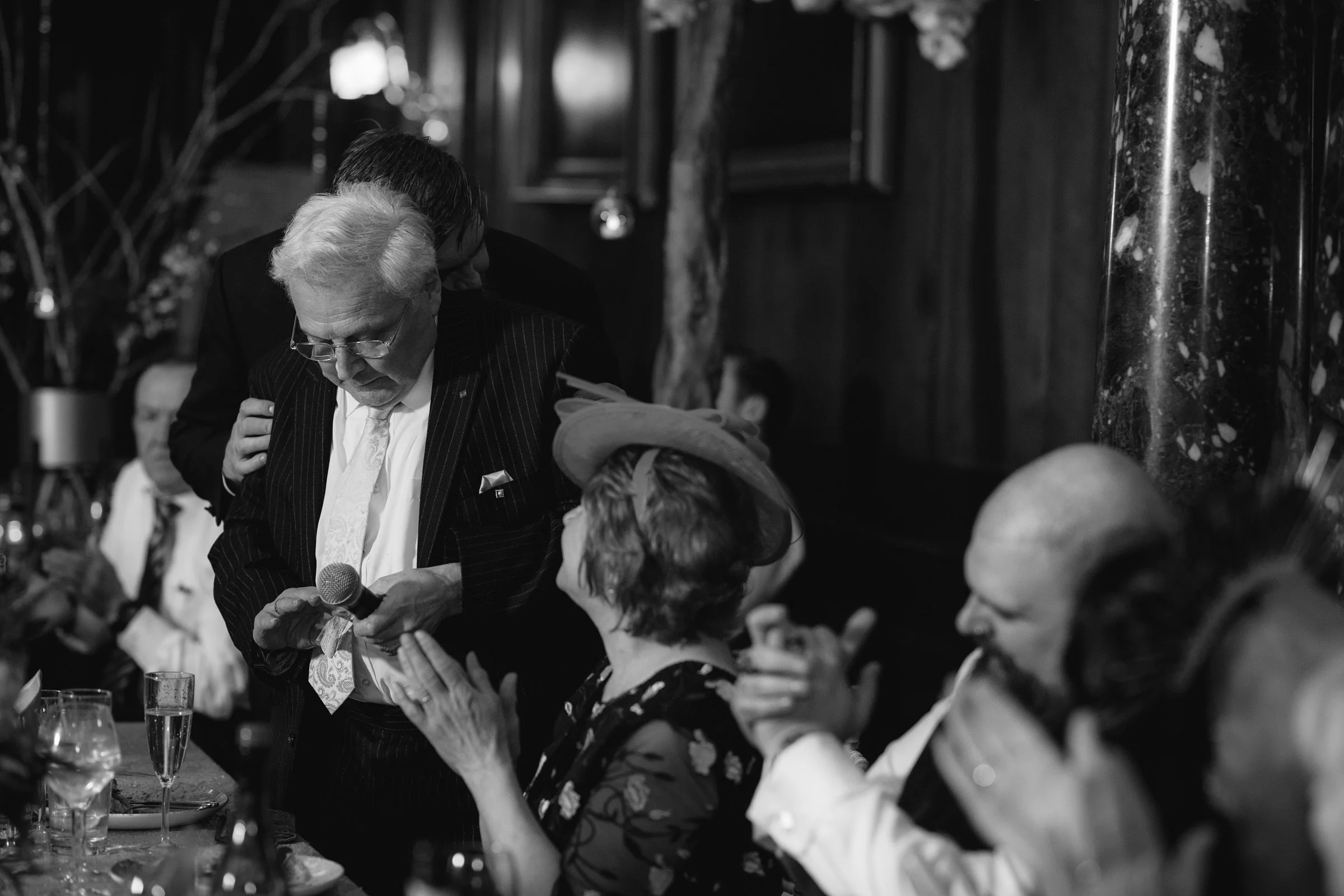 An elderly man in a suit holding a microphone appears to be giving a speech at a formal gathering. He is surrounded by seated guests, including a woman wearing a hat and a man with a bald head. The scene is in a dimly lit, elegant setting, possibly a
