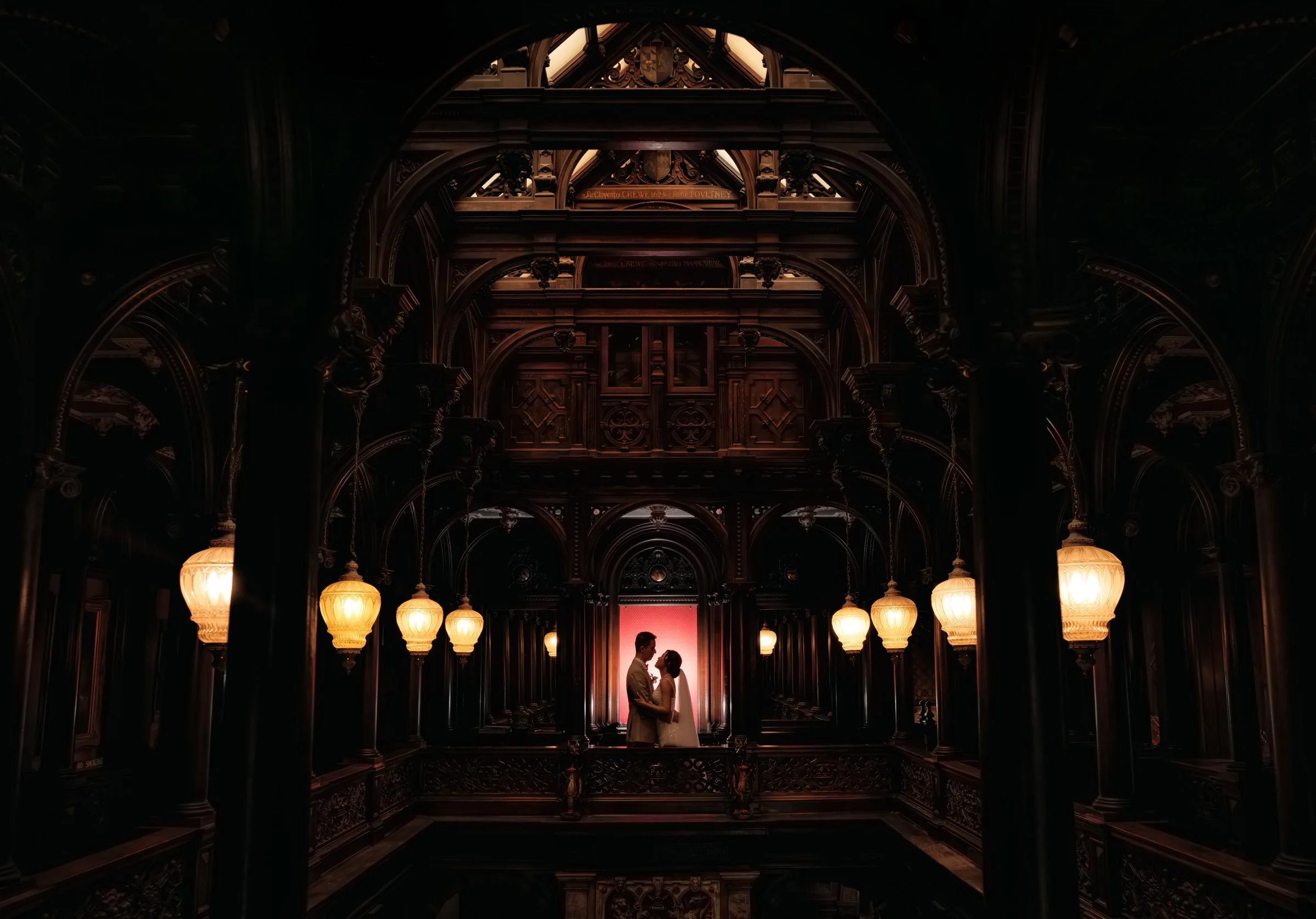 A romantic scene of a man and woman in  wedding attire standing close together, facing each other, inside an ornate, dark wood hall with decorative archways, warm hanging lamps, and a pink backdrop.
