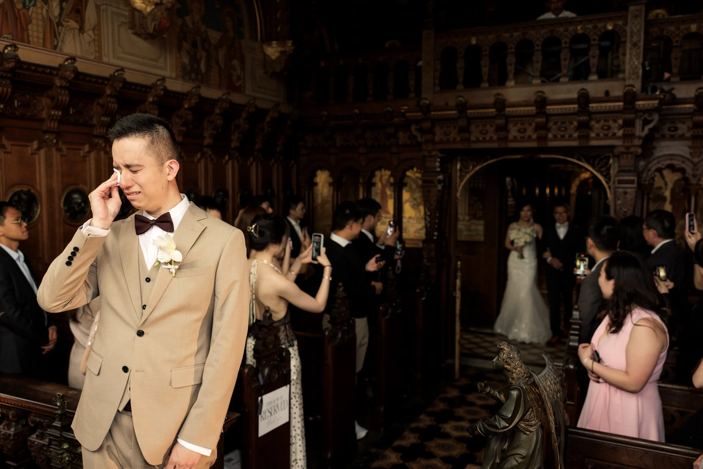 A young man in a tan suit and bow tie appears emotional, wiping away tears in a crowded room with ornate wood-paneled walls and multiple people, some taking photos, in formal attire.