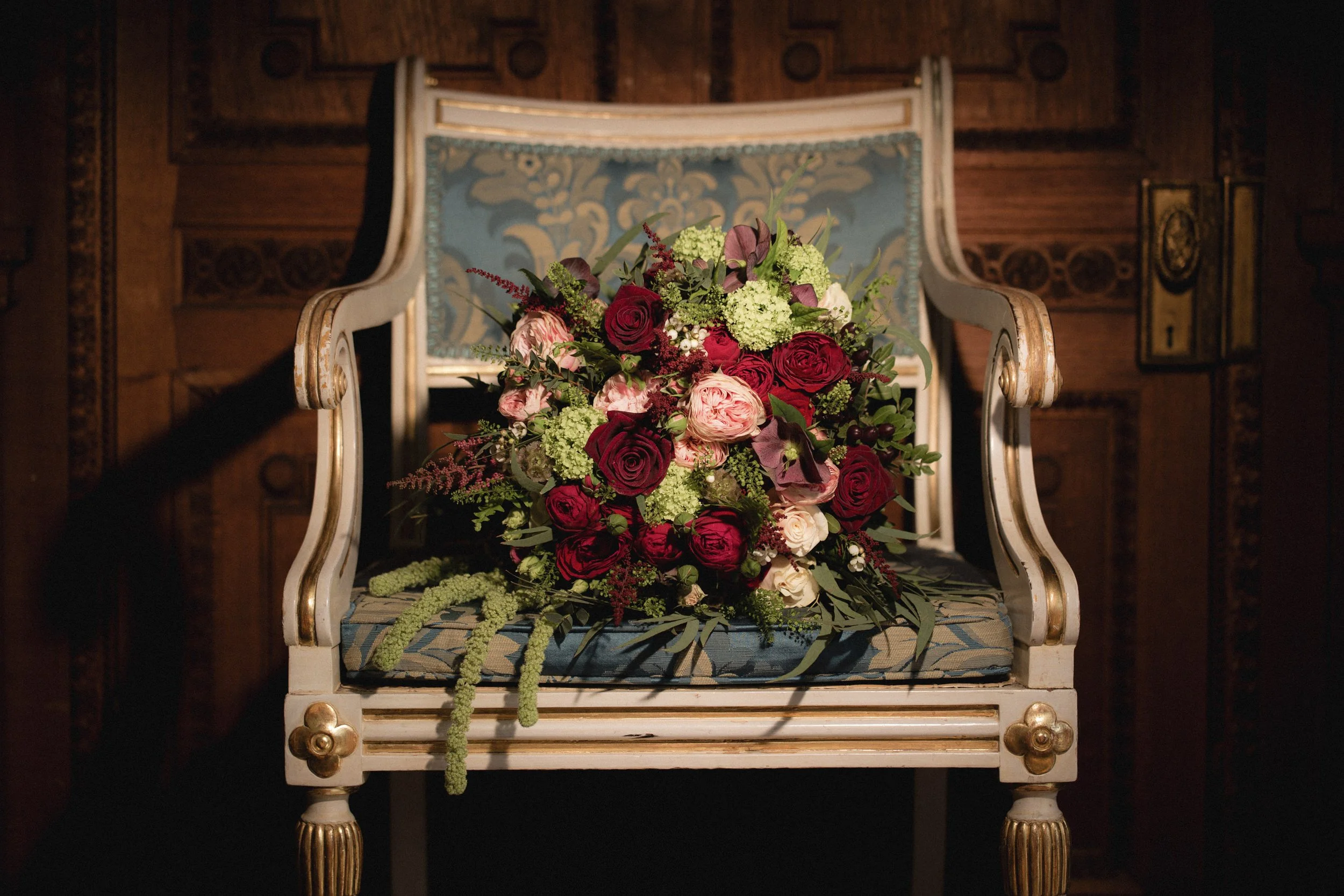 A bouquet of red, pink, white, and green flowers resting on an ornate blue and white vintage armchair with wooden carvings in a dimly lit room.